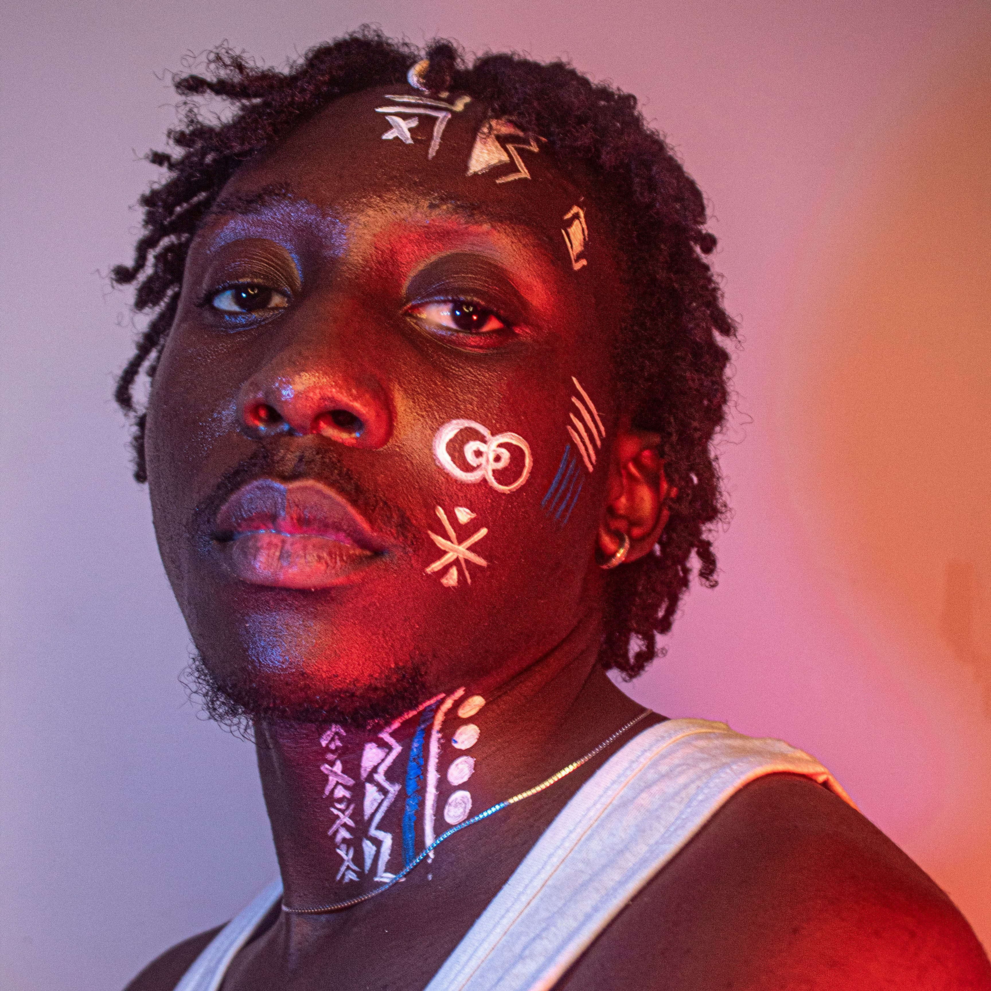 A man with traditional white face paint, lit in warm pink light, looking into the camera