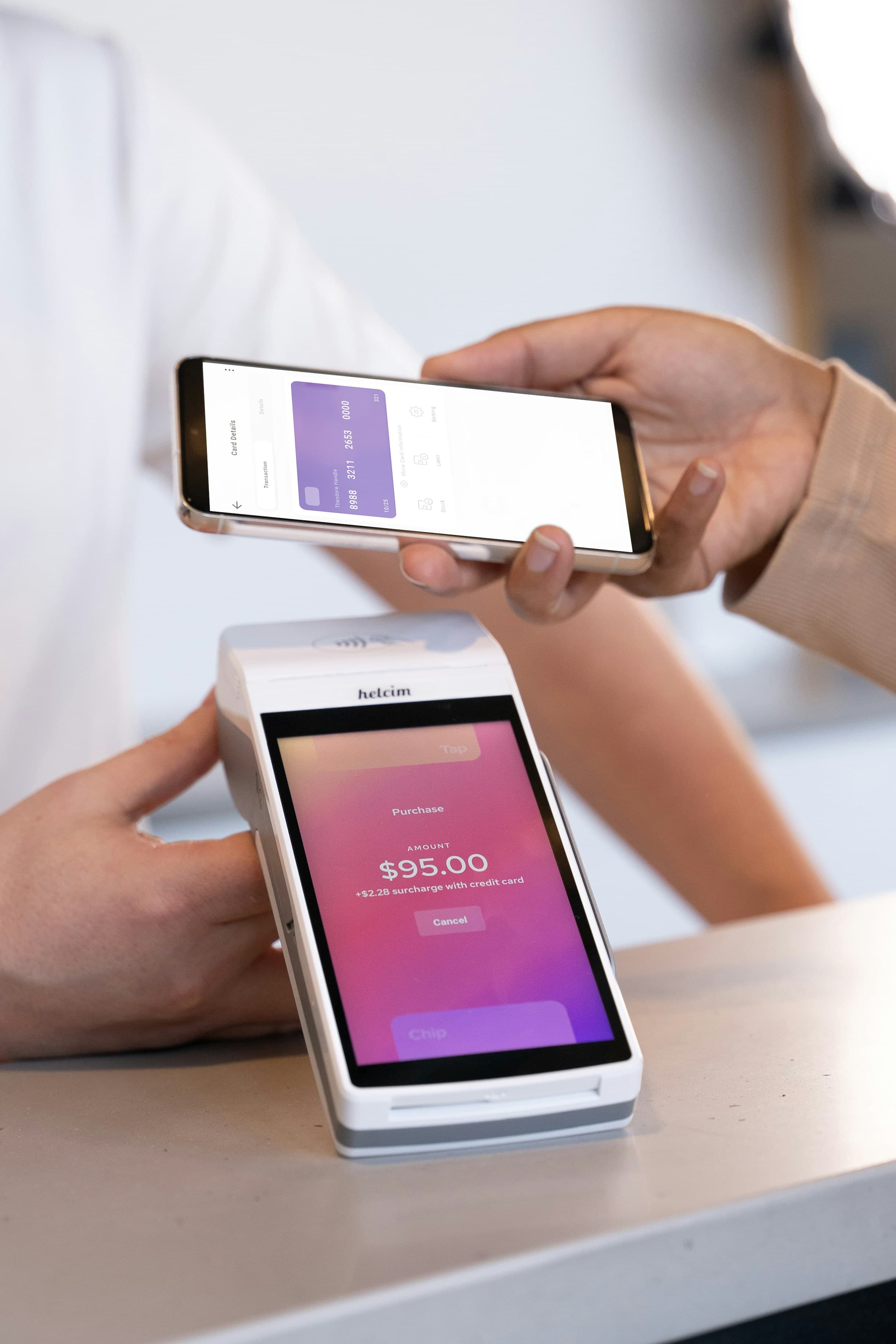 Hands holding a phone showing a purple bank card, tapping it on a payment terminal