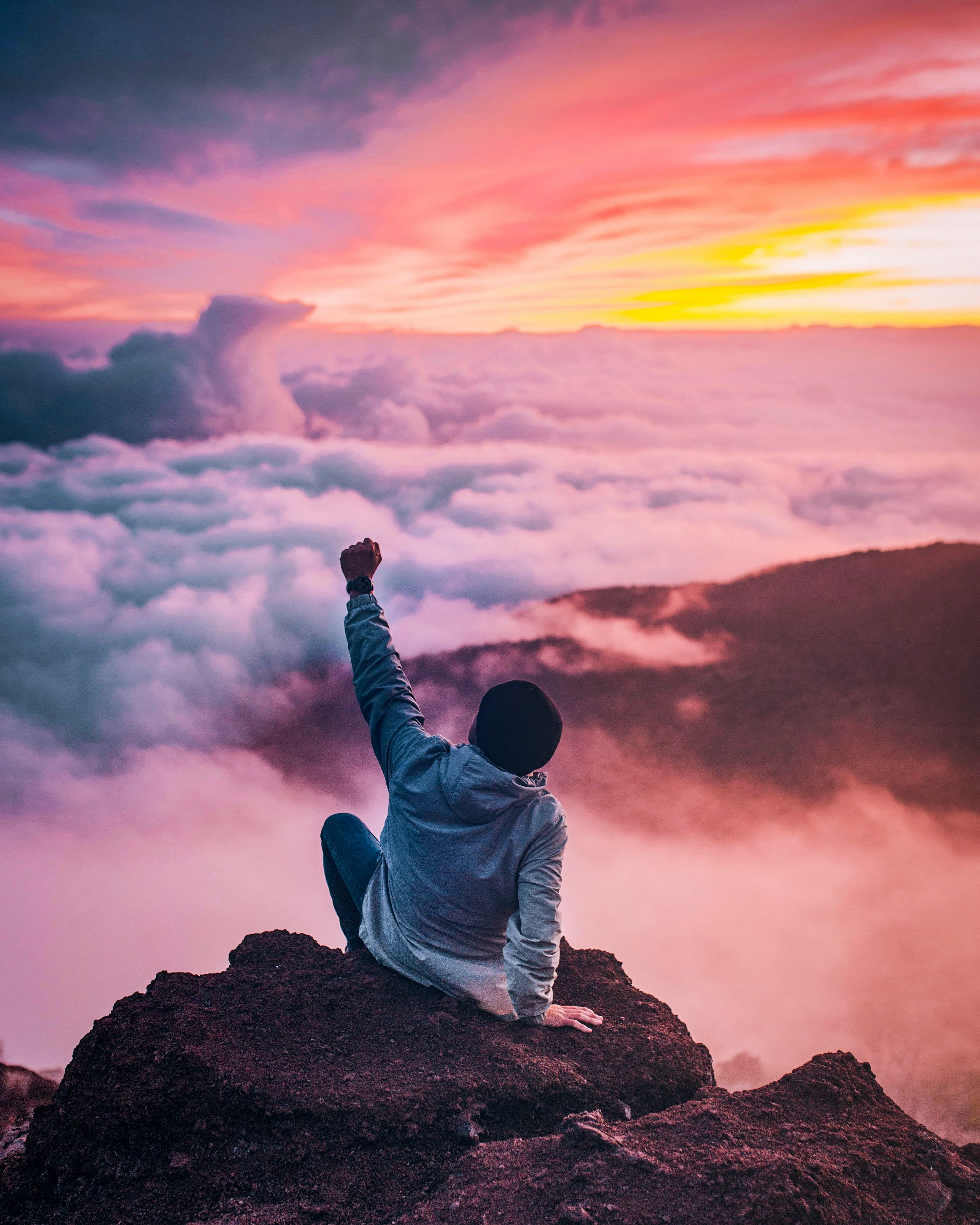 A person sitting on a mountaintop above the clouds at sunset, one fist raised